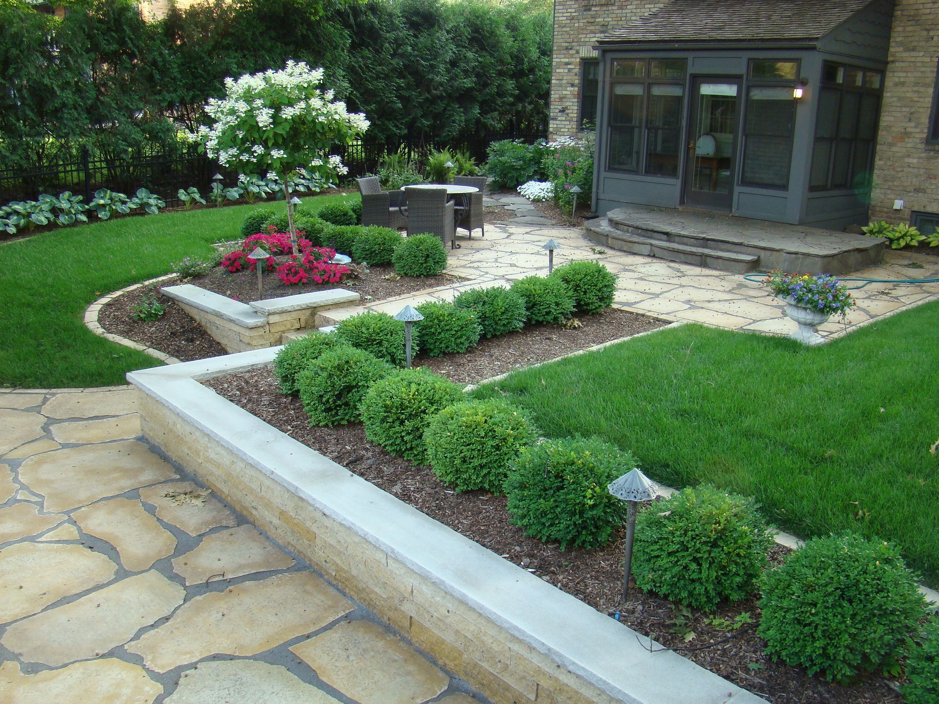 Stone patio with manicured lawn, shrubs, and flower beds near a house with a screened porch.