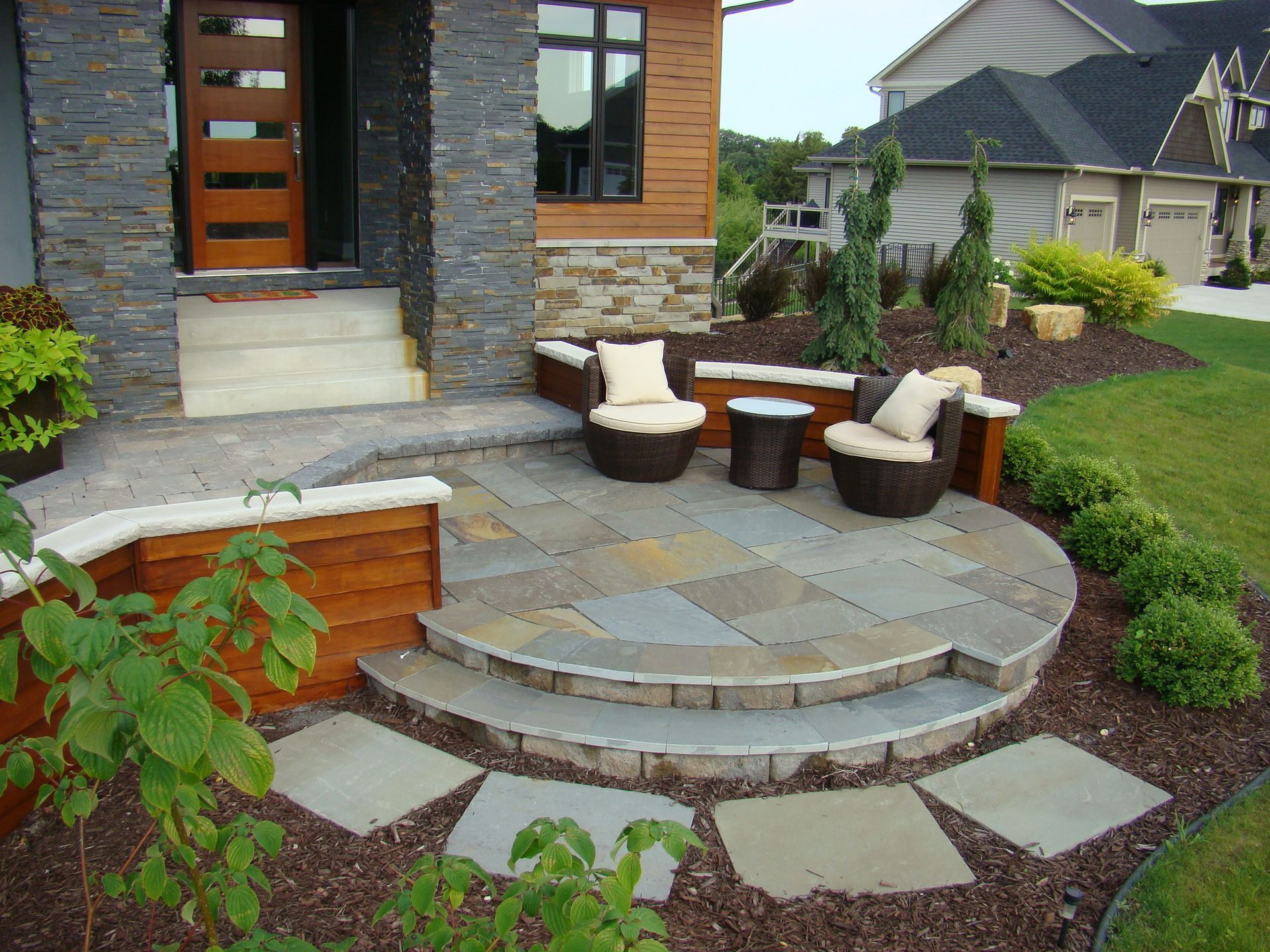 Patio with stone pavers, seating, and landscaping at a house entrance.