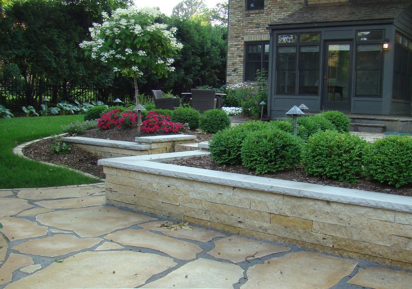 Stone patio with retaining walls holding shrubs and flowers, next to a house with a sunroom.