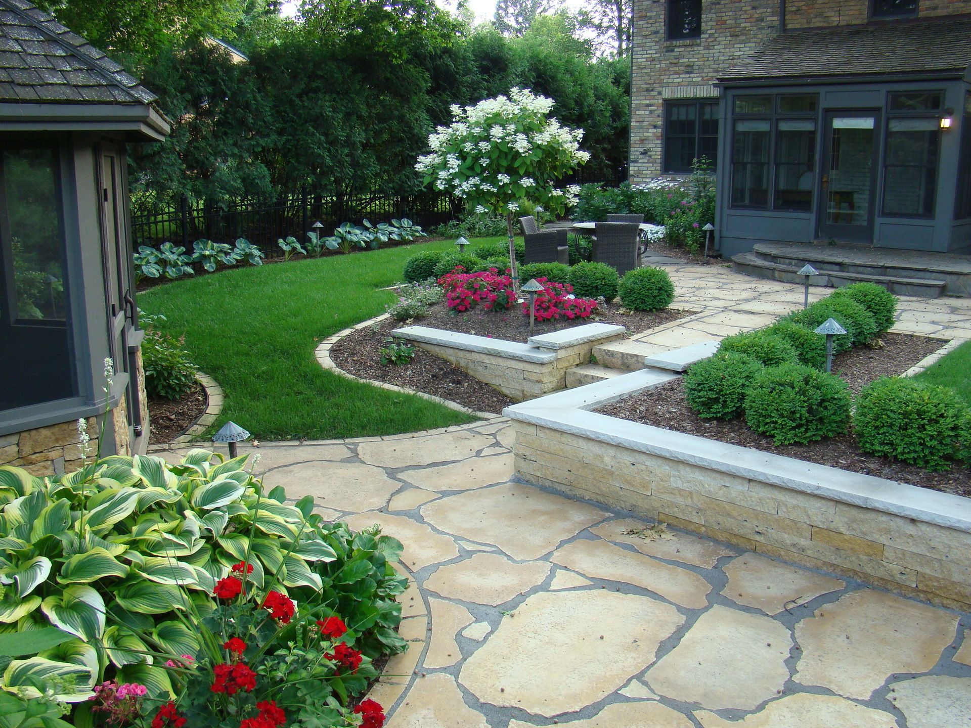 A stone patio with landscaped beds, green lawn, and a two-story house.