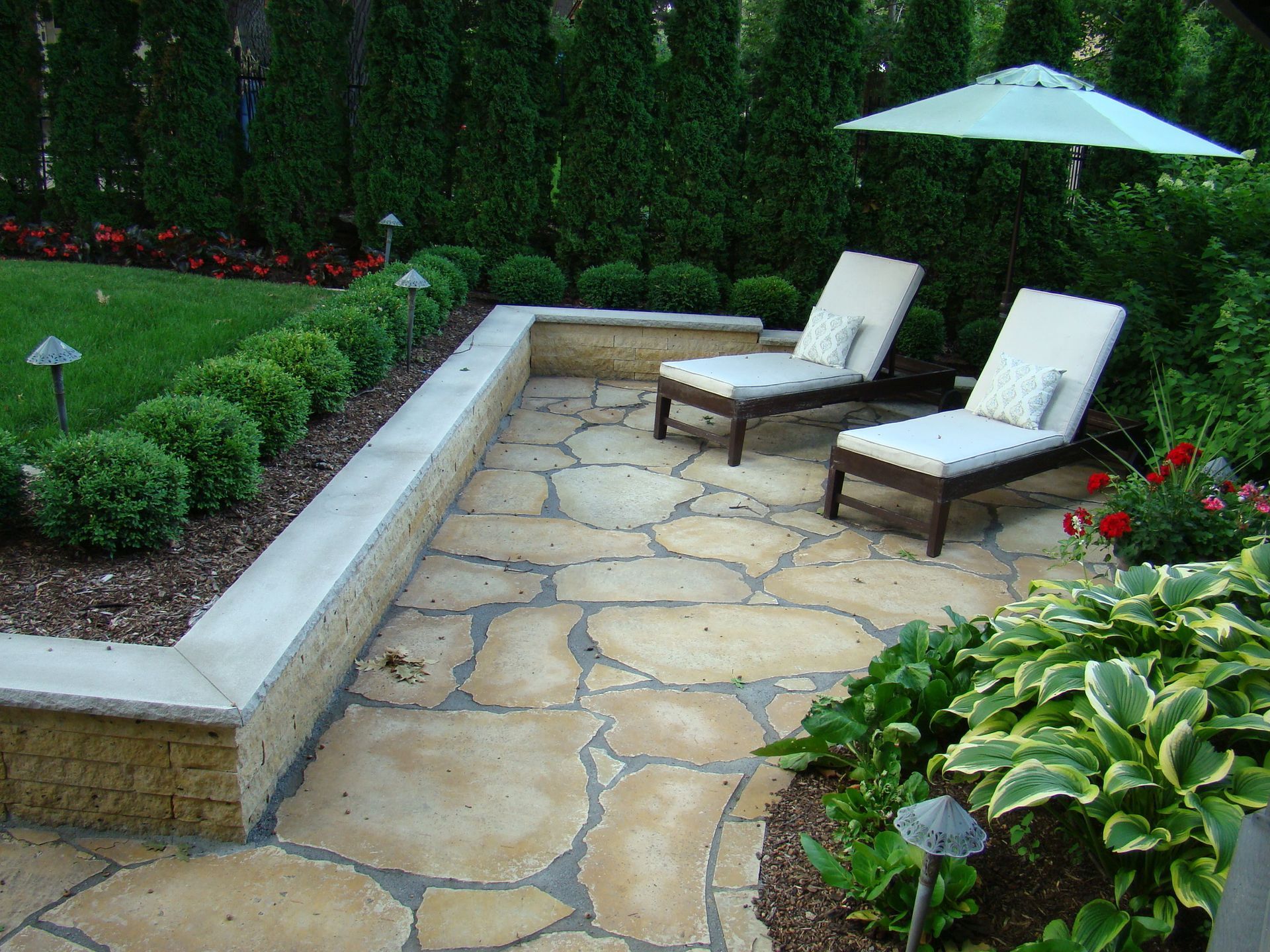 Stone patio with two lounge chairs, umbrella, and manicured landscaping, including bushes, flowers, and grass.