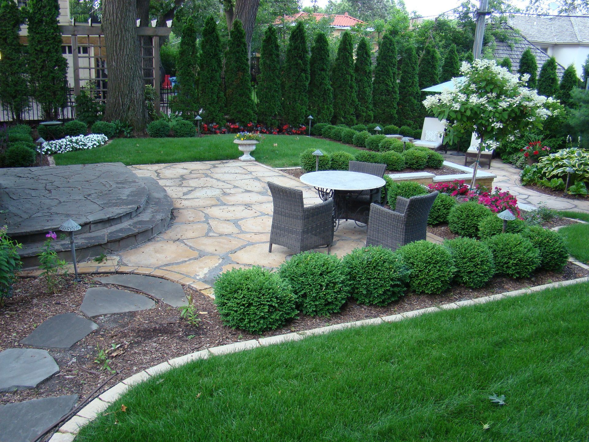 Stone patio with wicker chairs and table, surrounded by manicured shrubs and lawn.