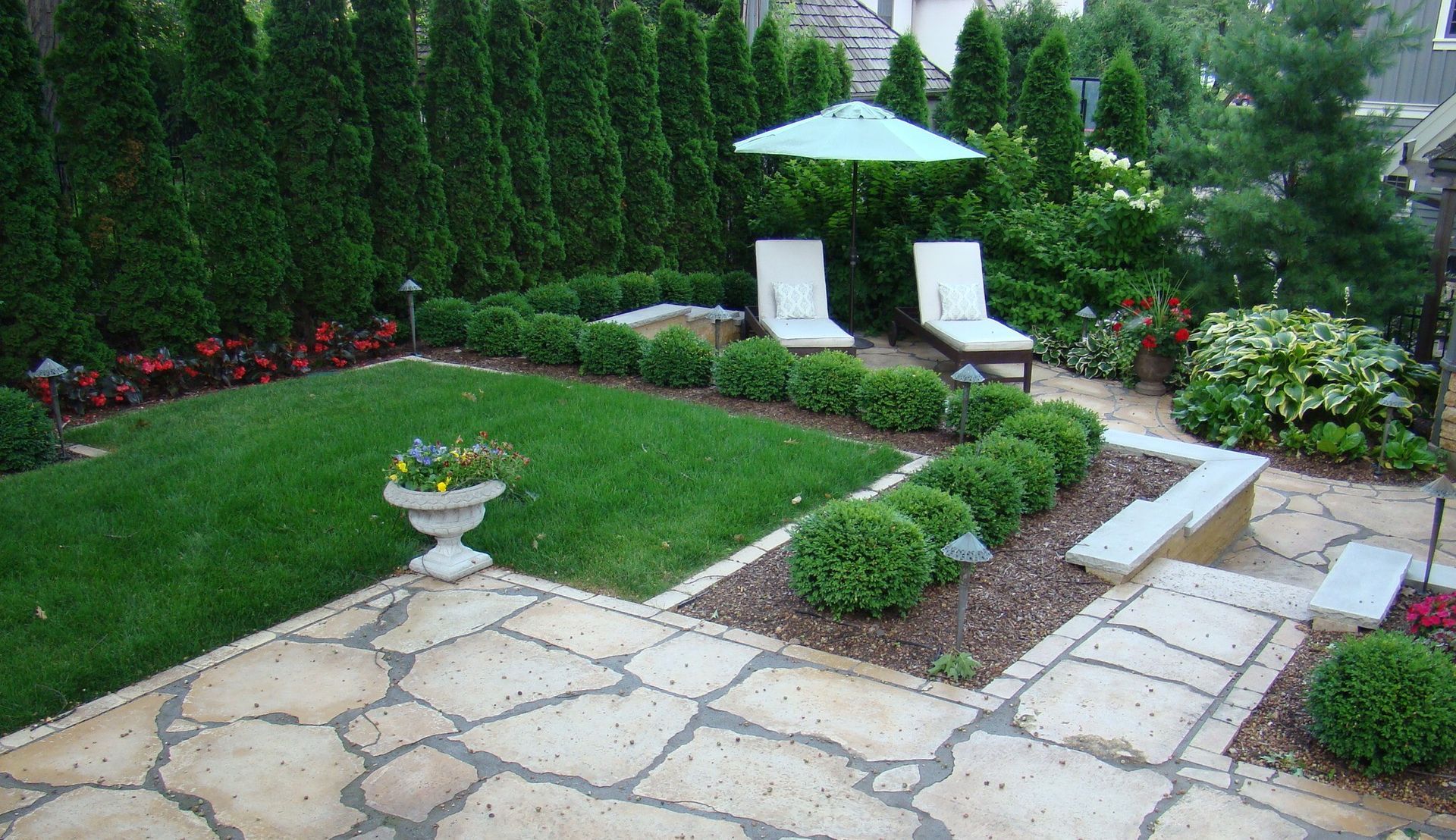 Lush green backyard with lawn, stone patio, shrubs, lounge chairs under an umbrella.
