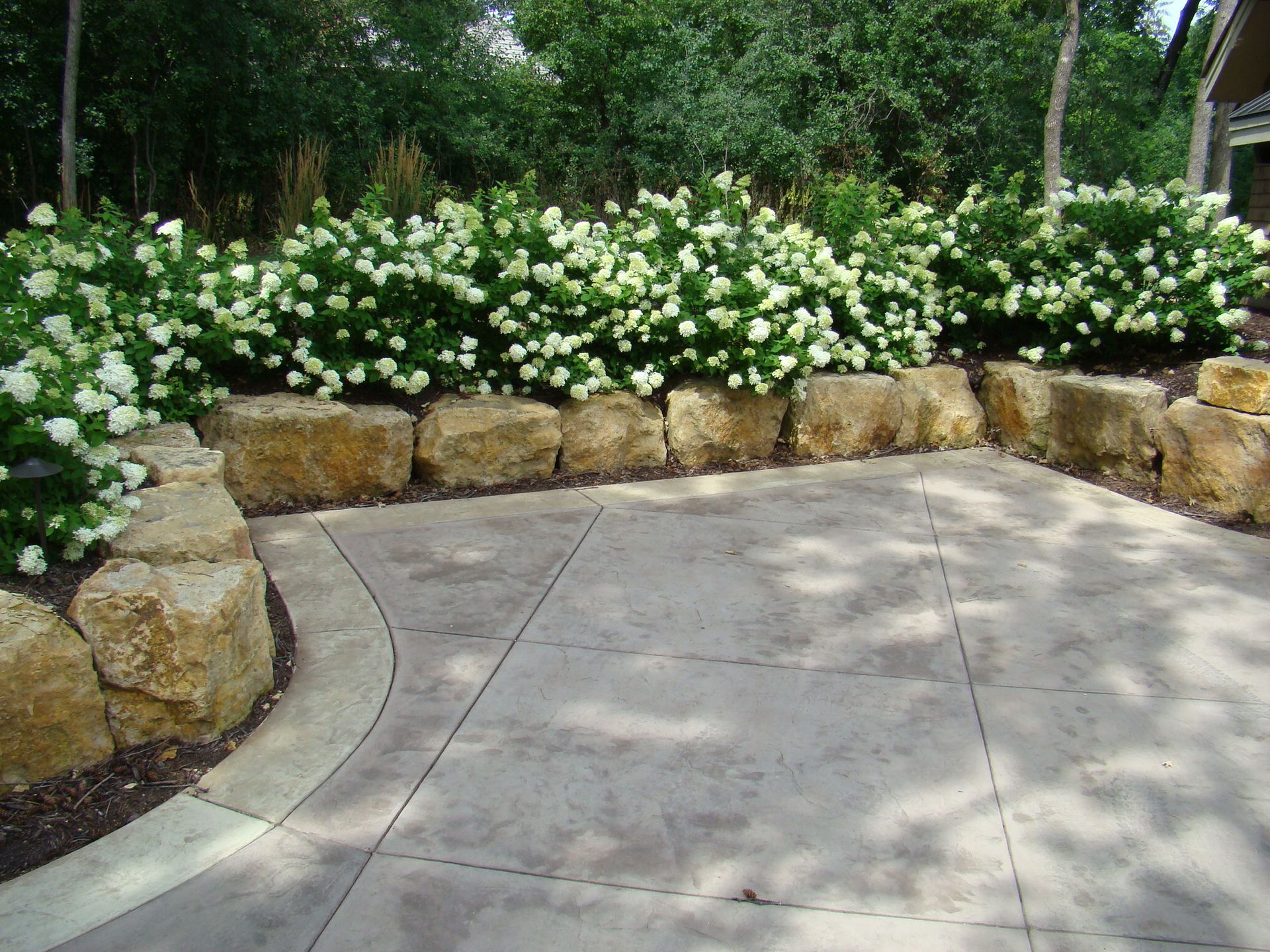 Patio with stone wall, white flowering shrubs, and trees in the background.