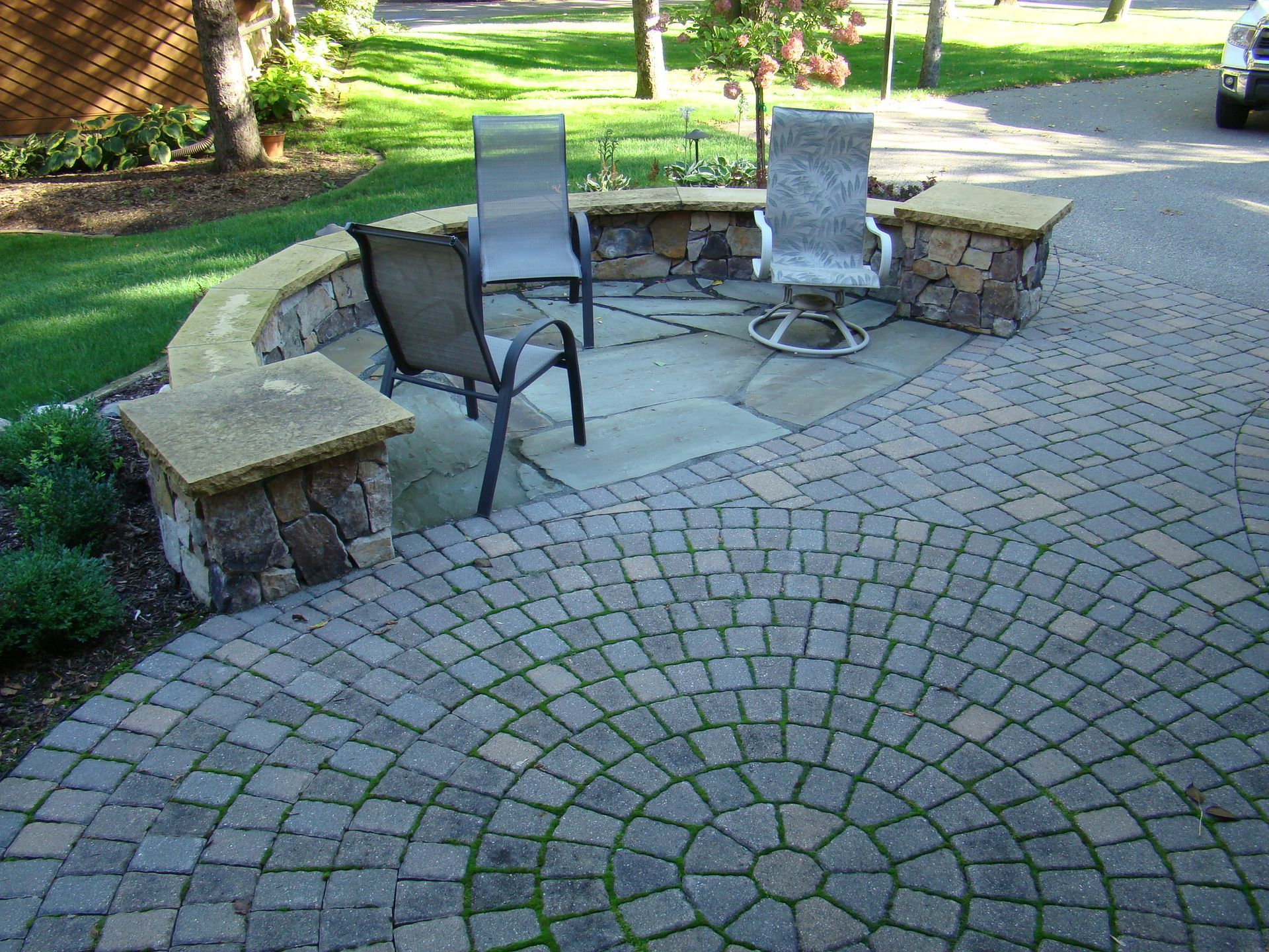 Stone patio with circular brick pattern, seating area with chairs, stone wall, and green grass.