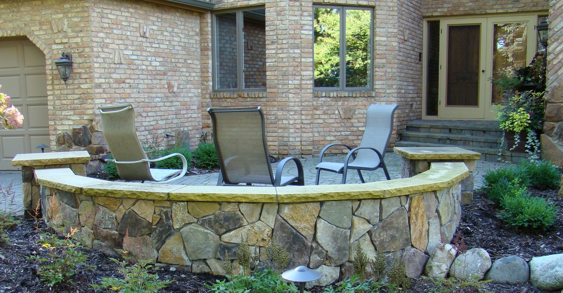 Stone-walled outdoor seating area with three chairs. Brick building and door in the background. Green bushes and grass around.