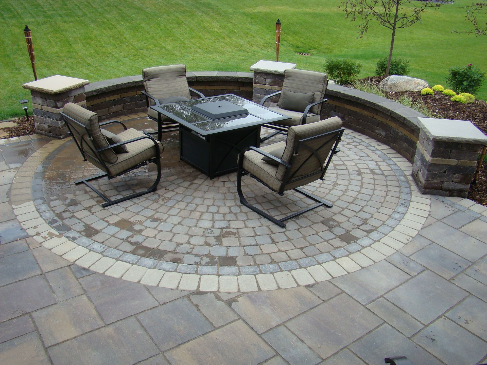 Patio with circular stone seating area, fire pit, and chairs. Low curved wall and green lawn in background.