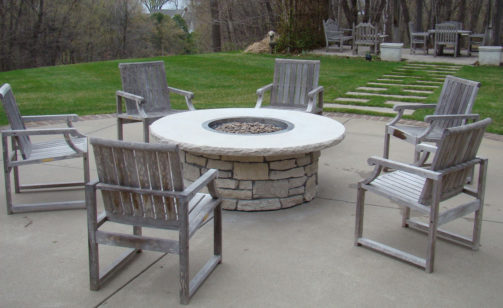 A circular stone fire pit surrounded by six wooden chairs on a patio; green grass in the background.
