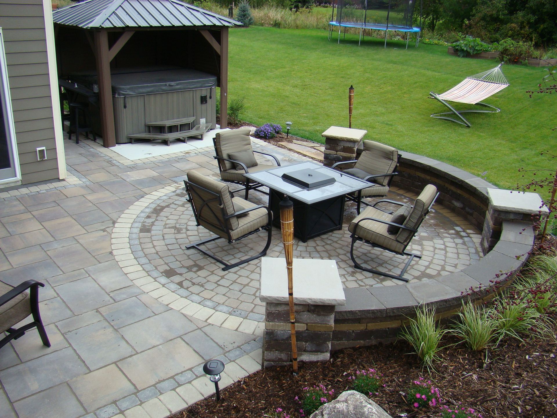 Backyard patio with fire pit, seating area, and hot tub under a gazebo, surrounded by grass.
