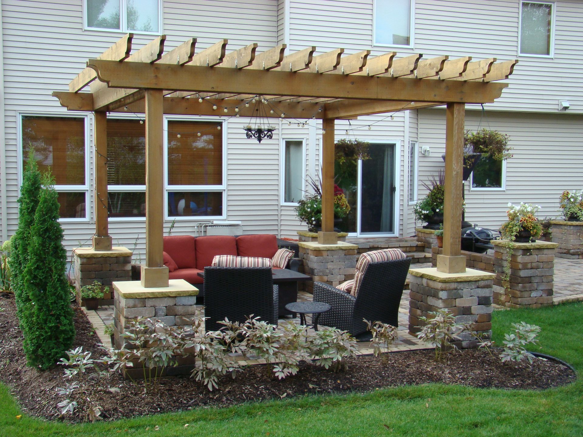 Pergola-covered patio with seating, built on stone columns, next to a house with windows and a sliding door.