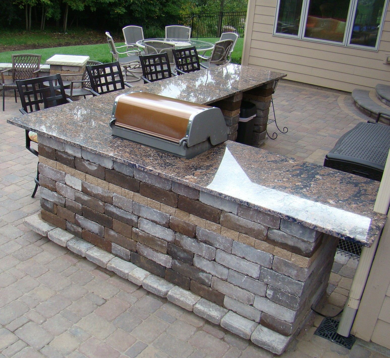 Outdoor kitchen with granite countertops, brick facade, and built-in grill. Patio seating visible.