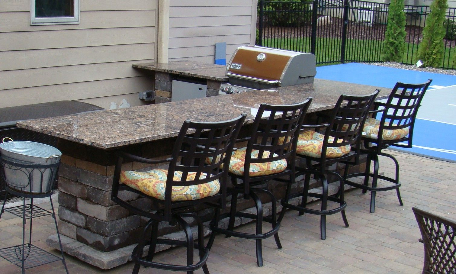 Outdoor bar with granite countertop, grill, and black bar stools with patterned cushions.