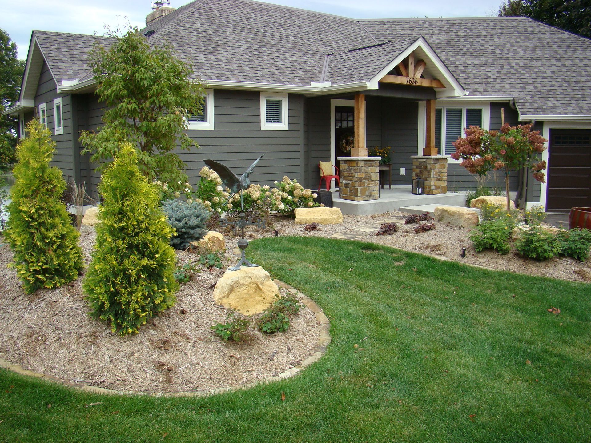 House with dark green siding, stone columns, and landscaped front yard with green lawn.