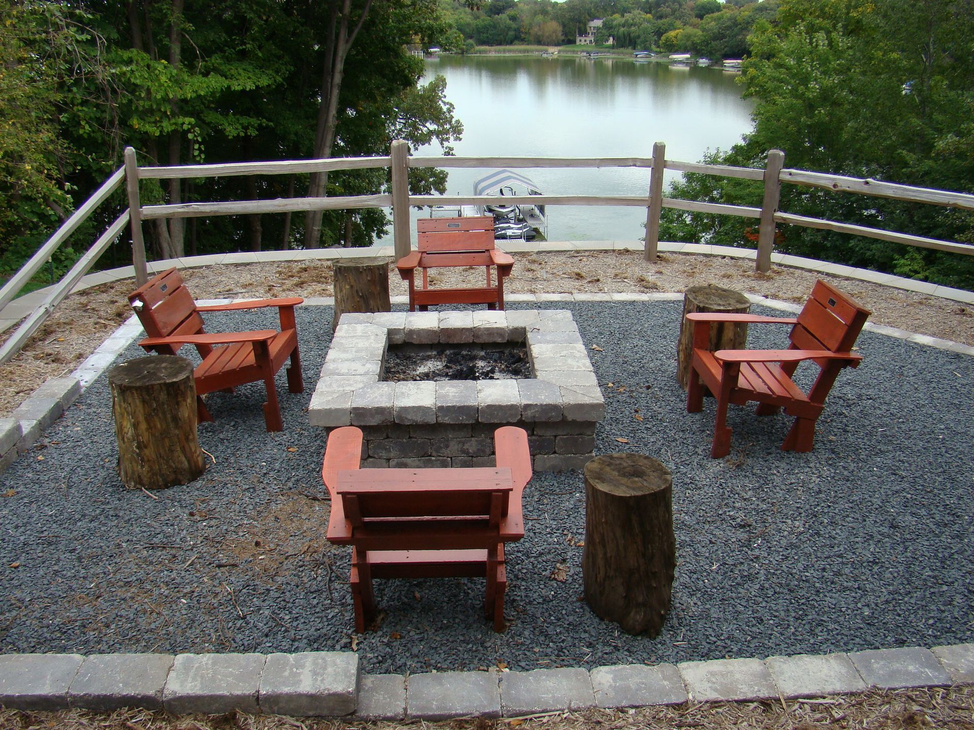 Fire pit with four red chairs and log stools, overlooking a lake.