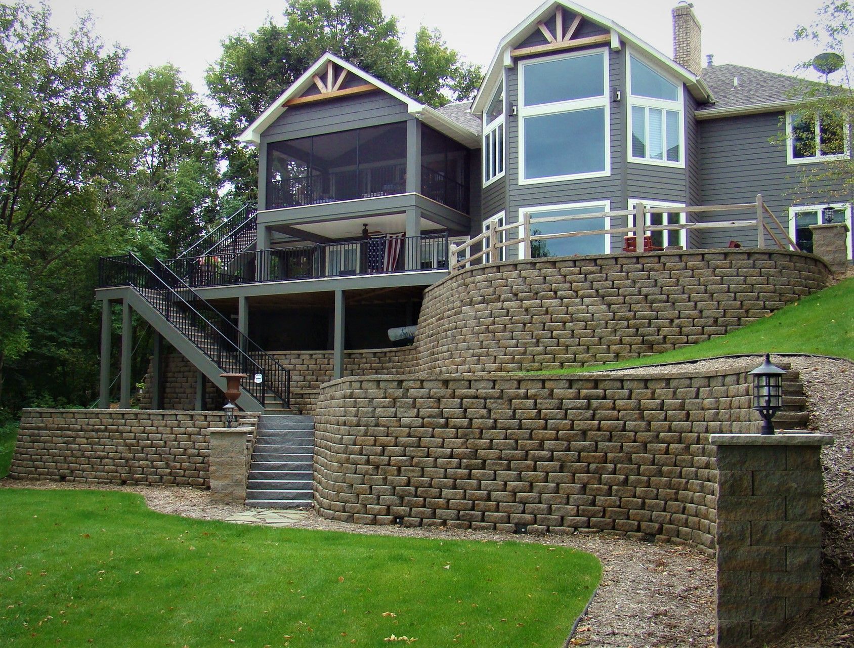 Grey house with tiered retaining walls, stairs, and a screened porch. Green lawn.