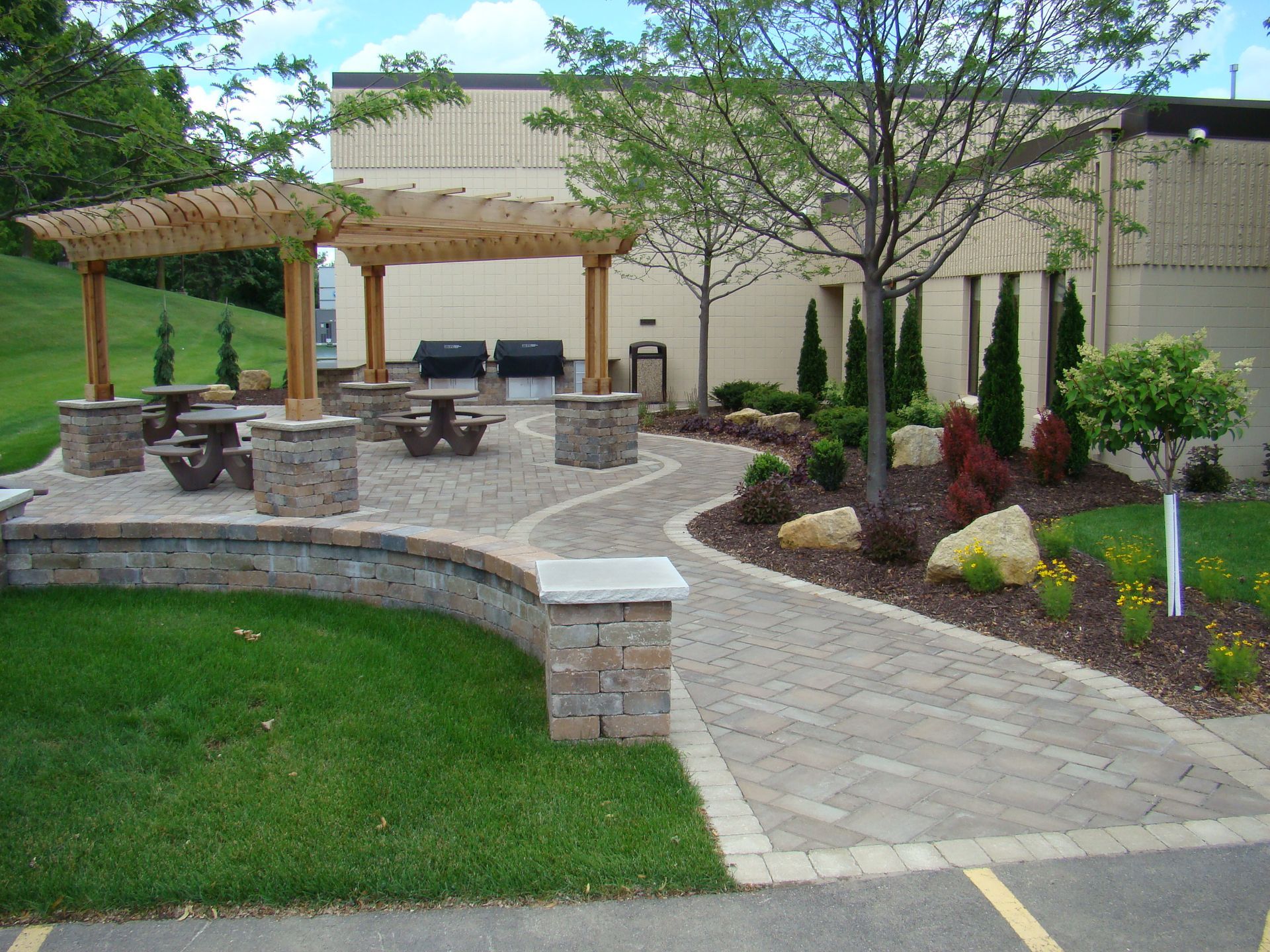 Patio with wooden pergola, picnic tables, and winding paved path surrounded by landscaping.