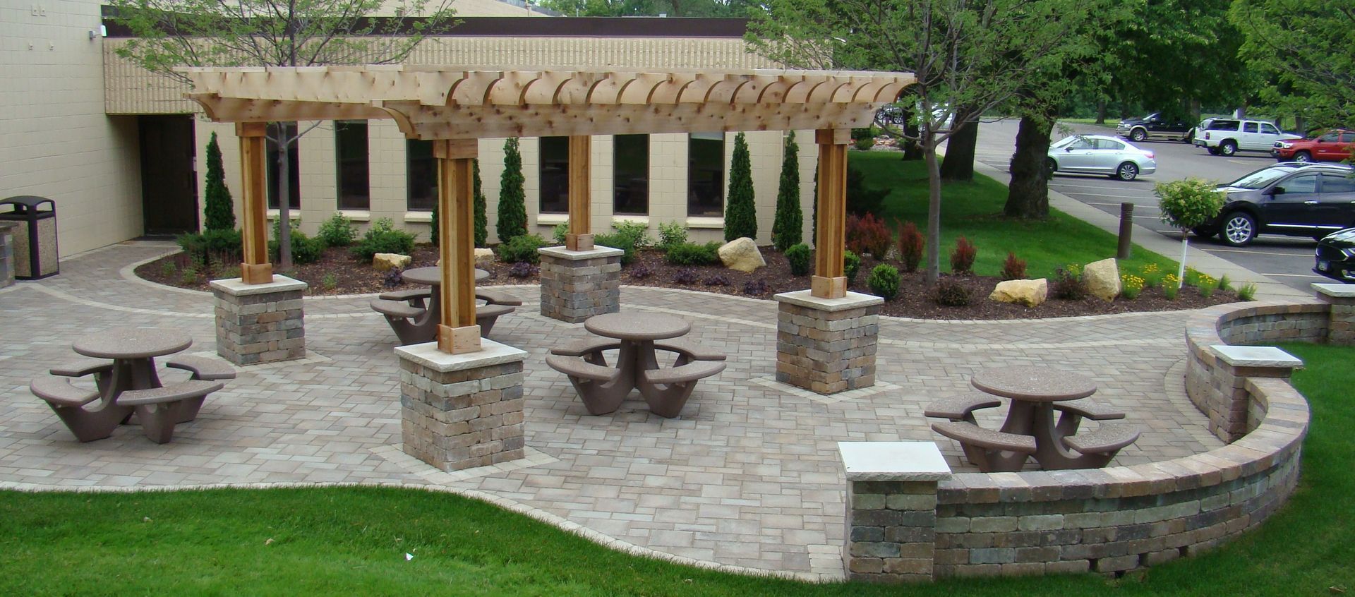 Outdoor patio with picnic tables under a wooden pergola, surrounded by greenery and stonework.