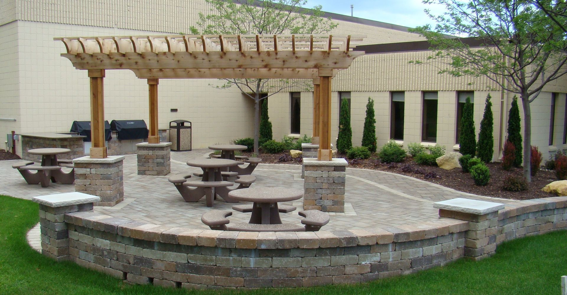 Outdoor patio with tables under a wooden pergola, surrounded by a low brick wall and landscaping.
