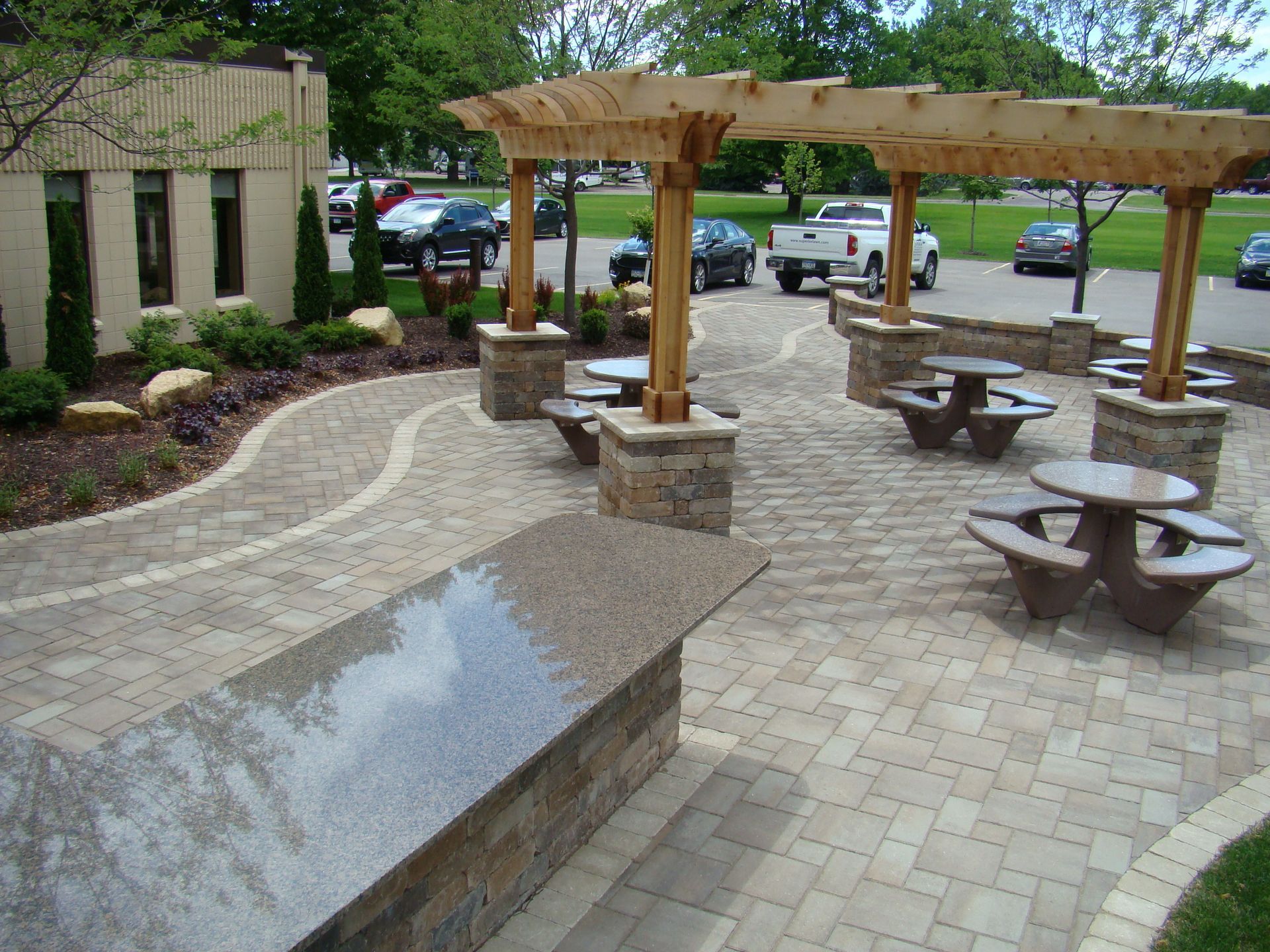 Outdoor patio with stone pavers, picnic tables under a wooden pergola, and a stone retaining wall.