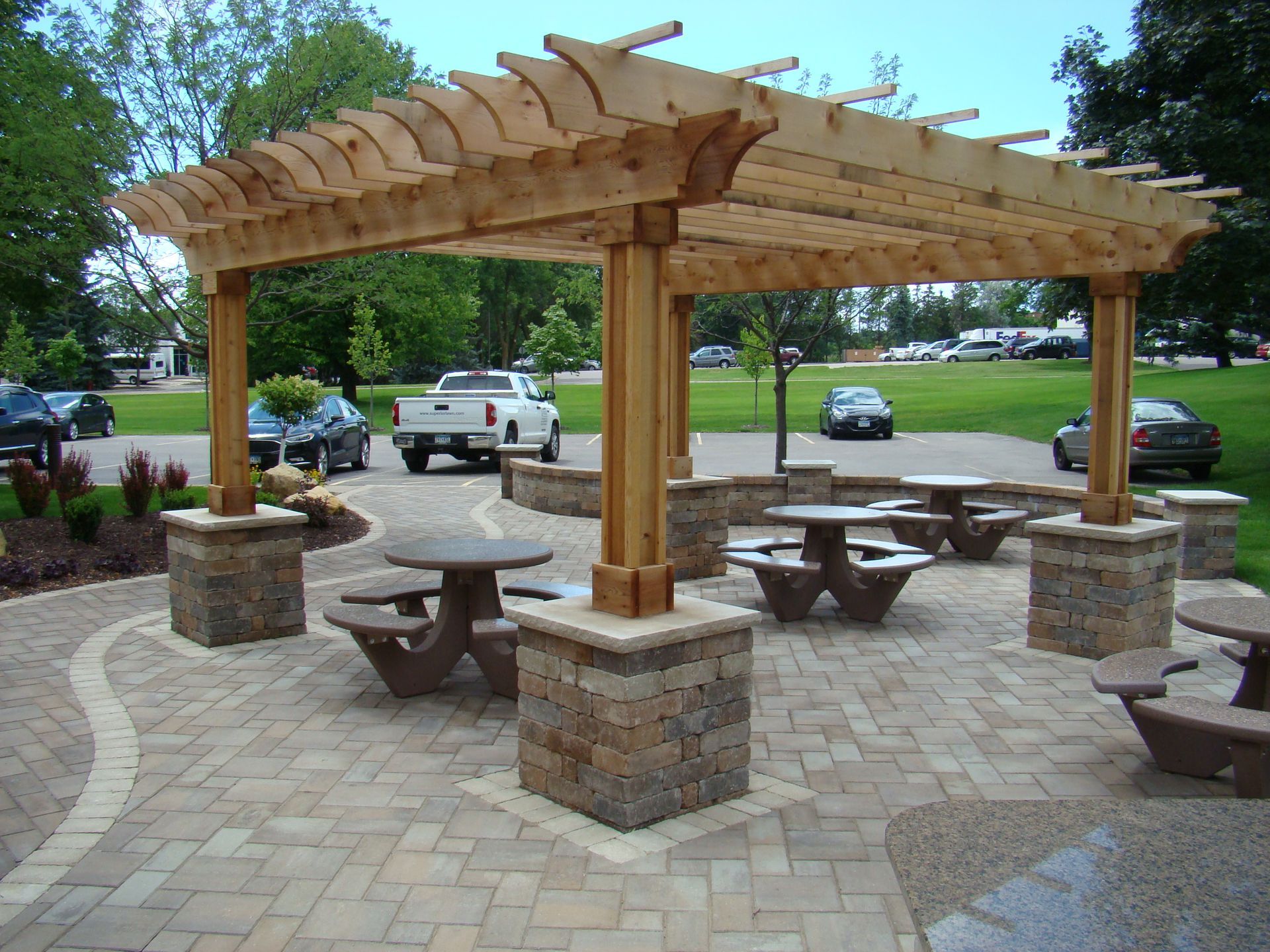 Wooden pergola over picnic tables on a brick patio in a park.