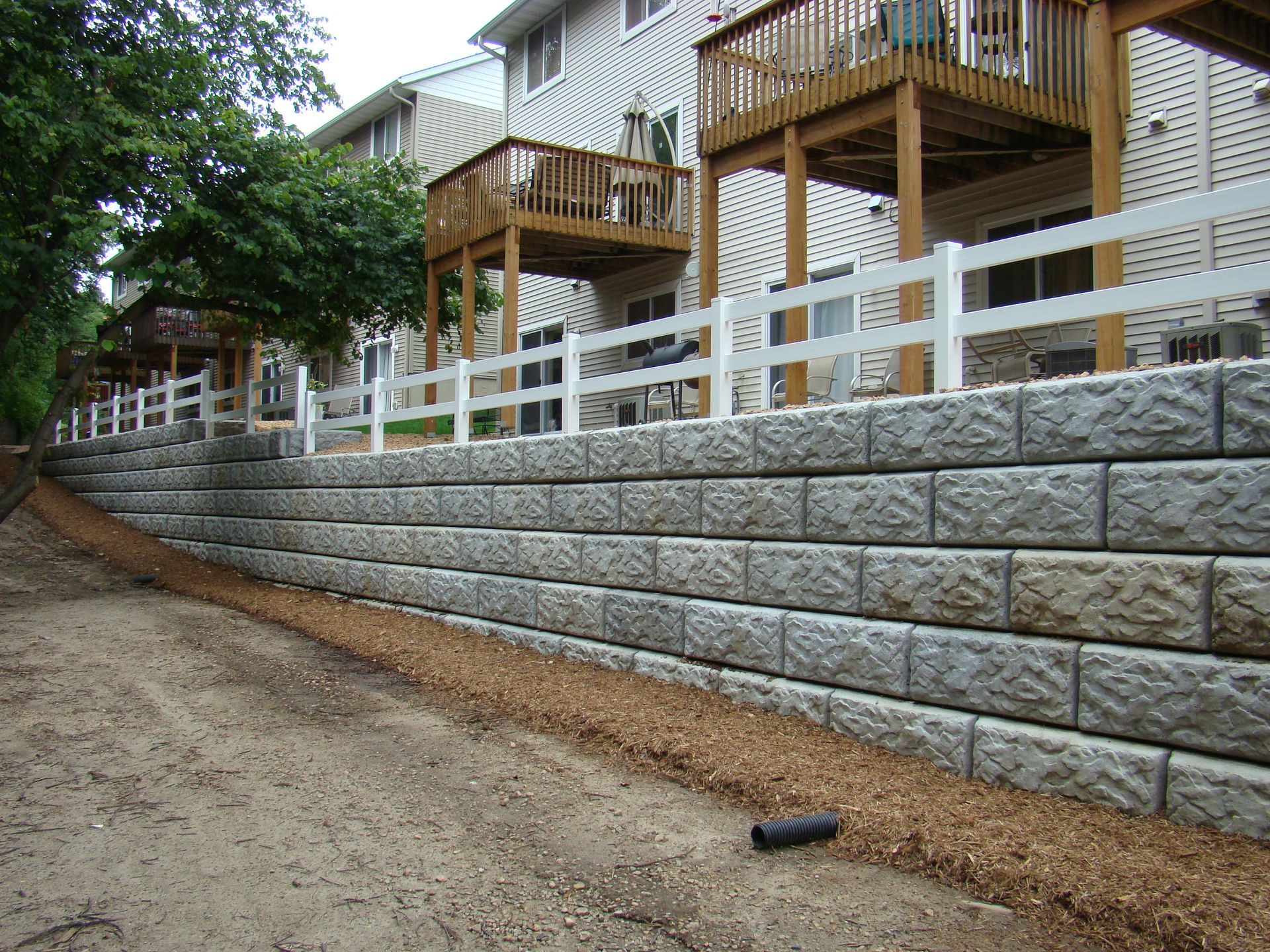 Retaining wall made of gray blocks with a white fence above. Houses and trees in background.