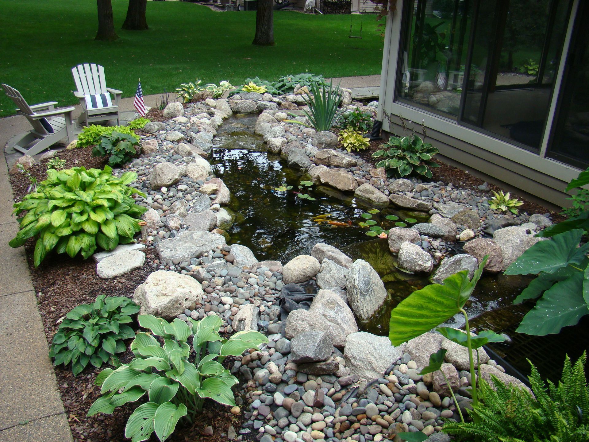 Pond with rocks and plants near a house, with chairs and a flag in the background.