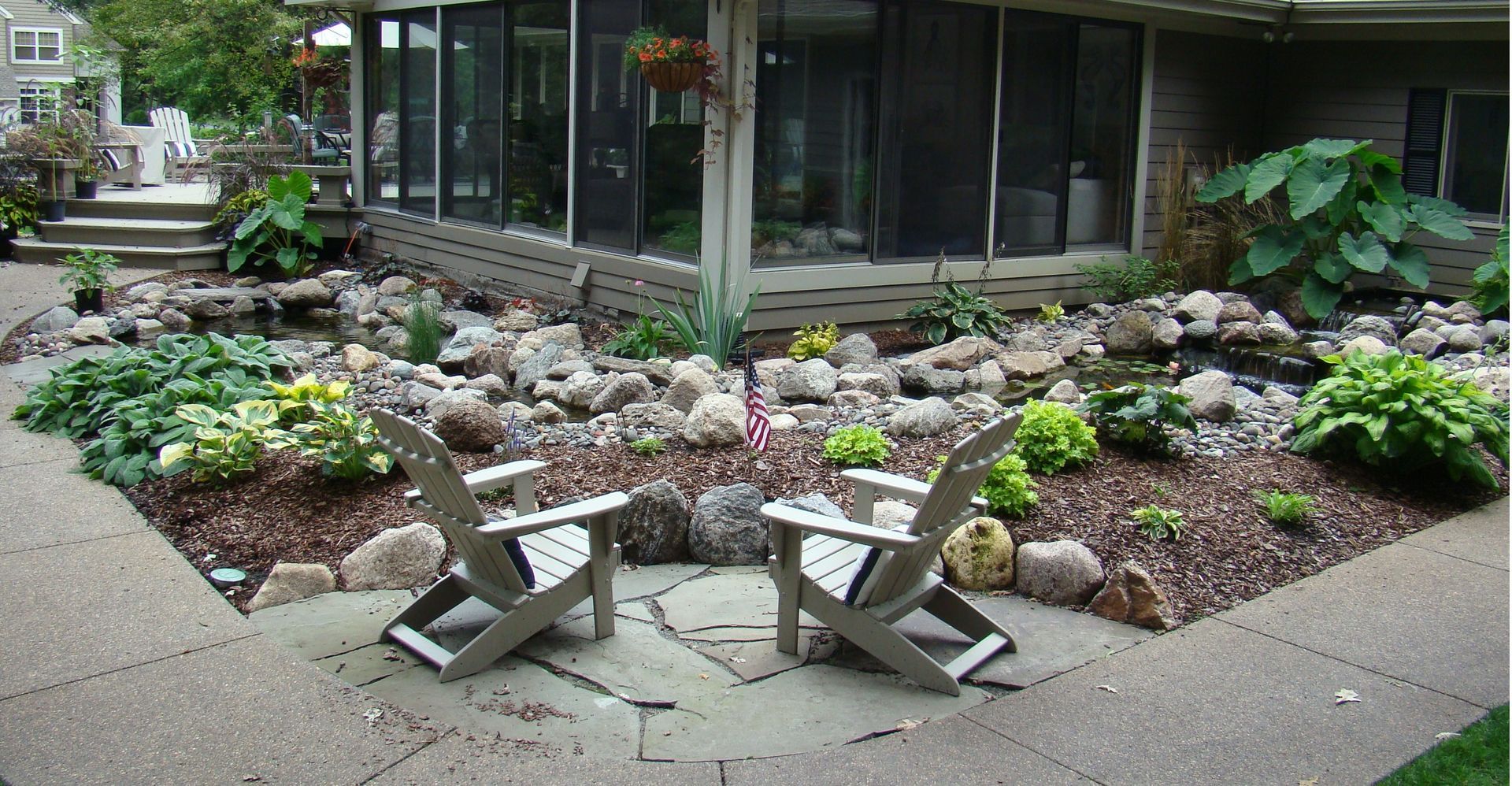Two wooden Adirondack chairs face a rock garden and pond near a building.