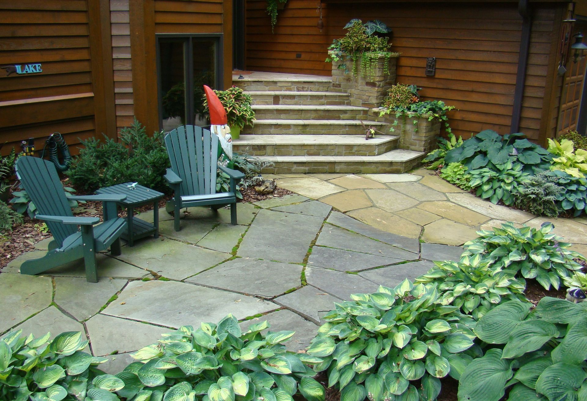 Stone patio with Adirondack chairs, garden and steps to a wooden building entrance.