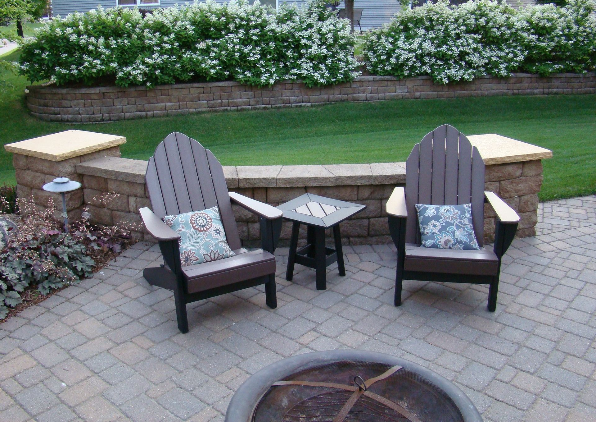 Two Adirondack chairs and a small table on a patio, with a brick wall and foliage in the background.