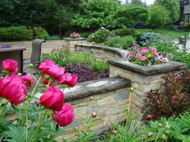 Stone garden beds with pink peonies and other flowers; trees in the background.