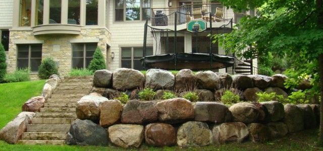 Stone retaining wall with steps leading up to a large house with a trampoline in the background. Green grass.