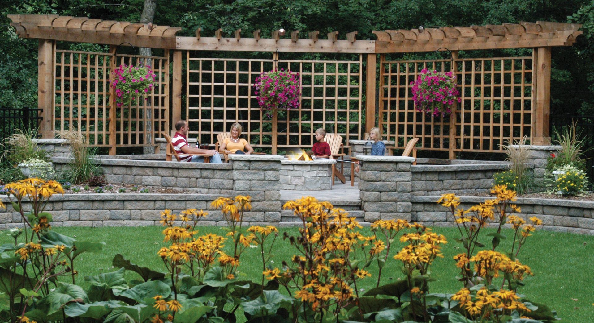 Outdoor seating area with pergola and stacked stone wall, flowers in foreground, people seated.