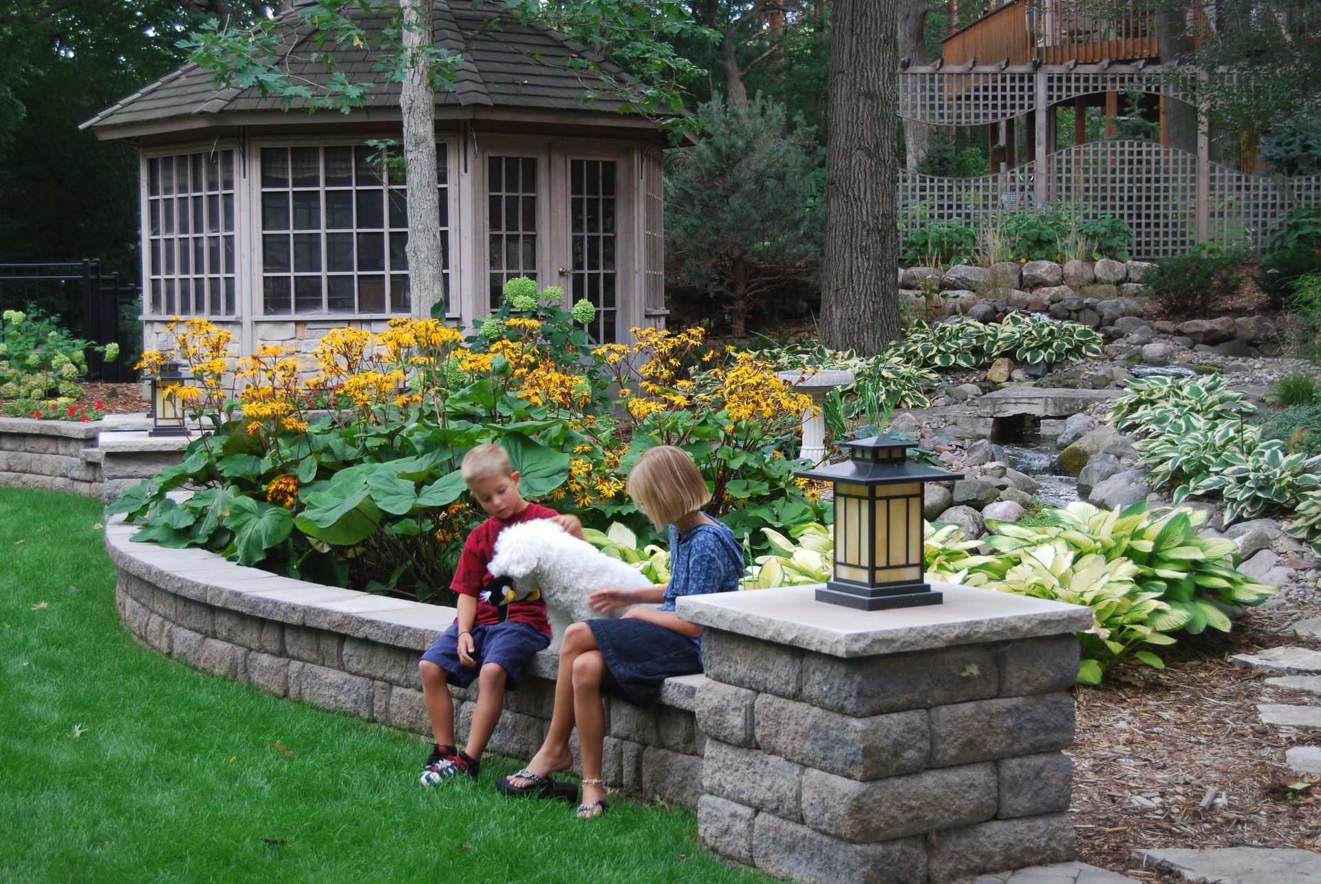 Two children and a dog sit on a stone wall overlooking a garden with a gazebo.