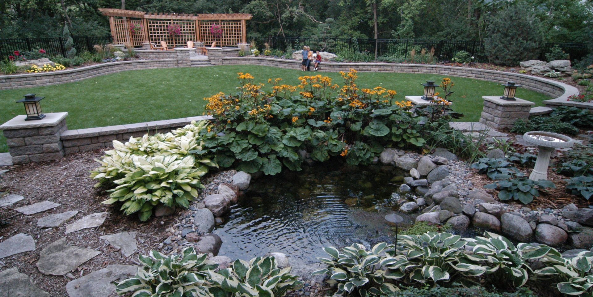 A manicured garden with a small pond, stone paths, and a grassy area, with a wooden structure in the background.