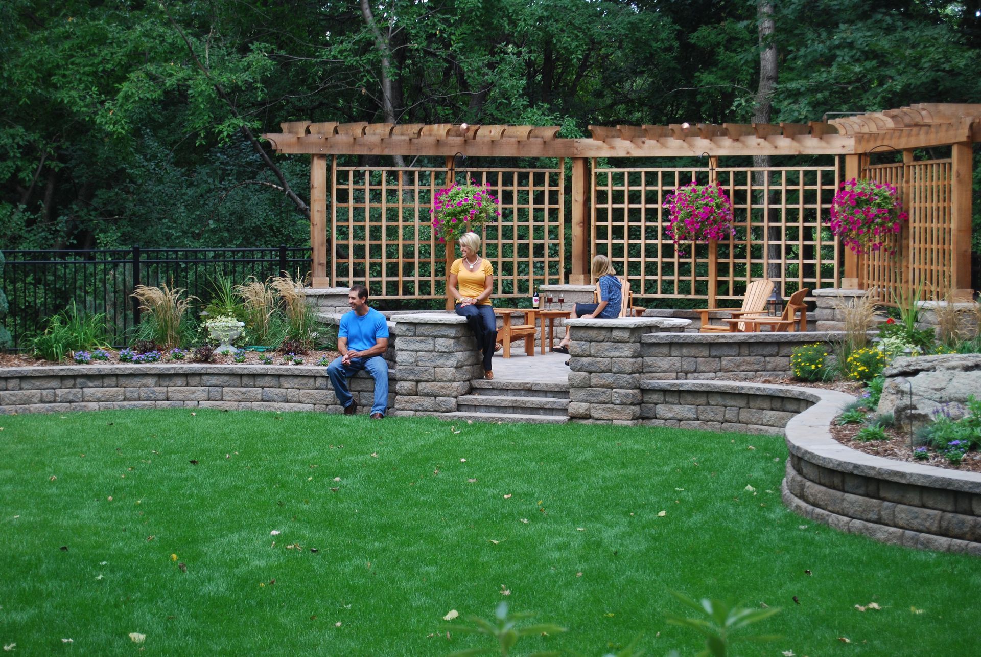 Lawn with a stone patio, trellis, and three people seated. Lush green yard, trees in the background, hanging flowers.