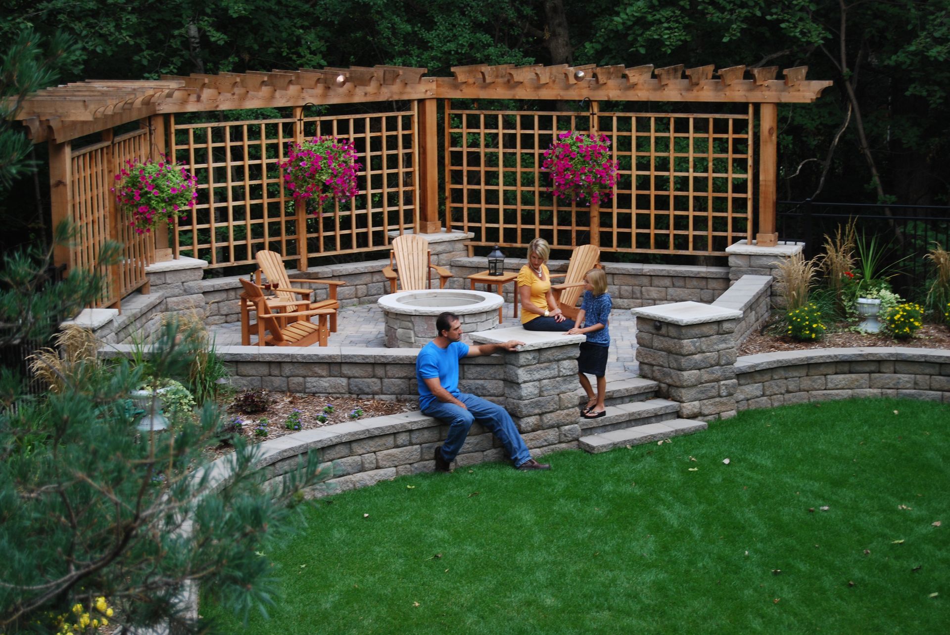 Outdoor patio with fire pit, wooden pergola, and people socializing. Lush green lawn and stone retaining walls.