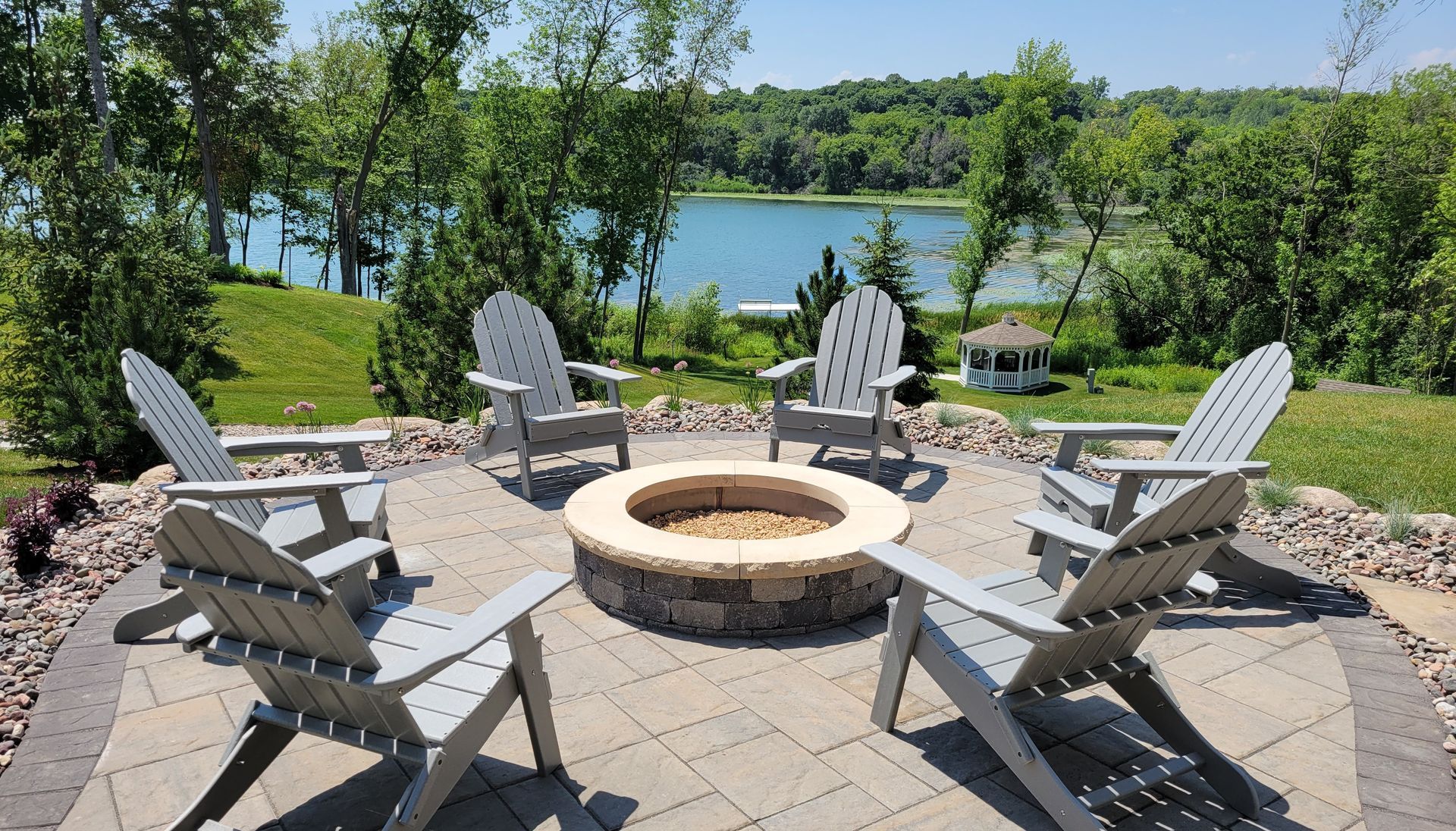 Six Adirondack chairs around a fire pit on a patio overlooking a lake.