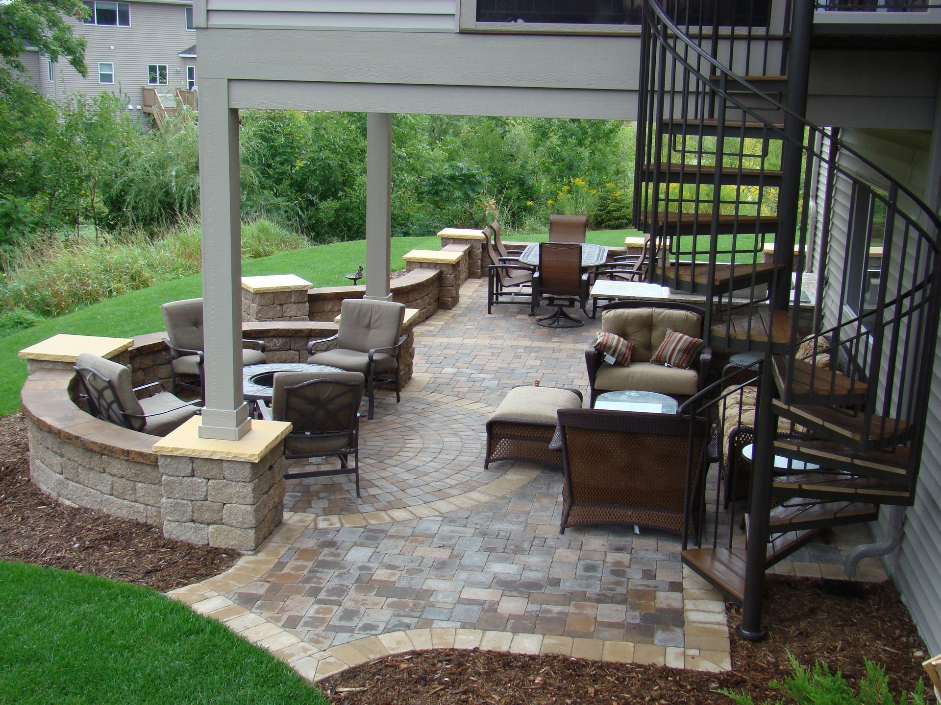 Patio with stone pavers, seating areas, spiral staircase, and lush greenery.