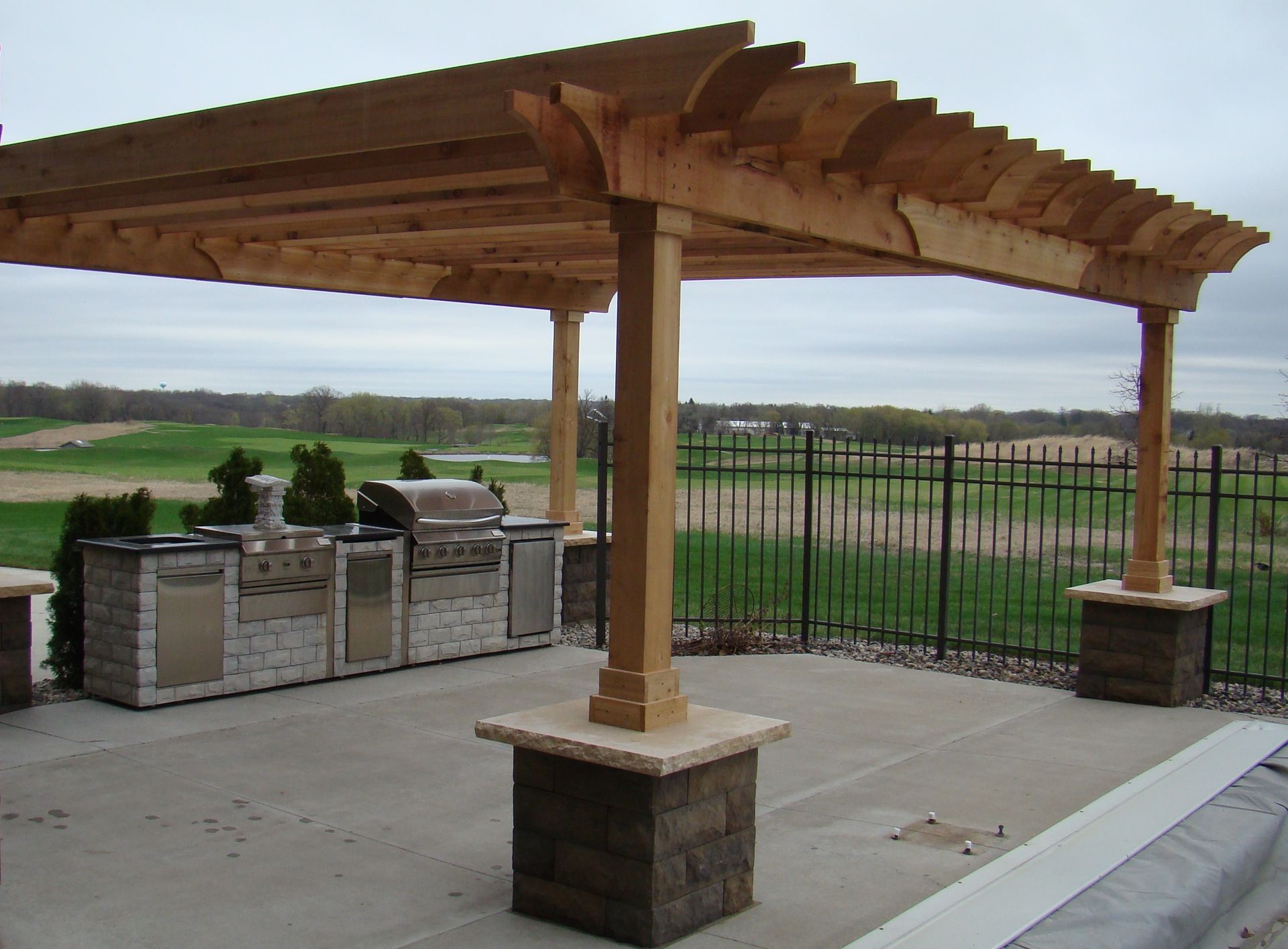 Wooden pergola over outdoor kitchen with grill and countertops. Concrete patio and view of a golf course.