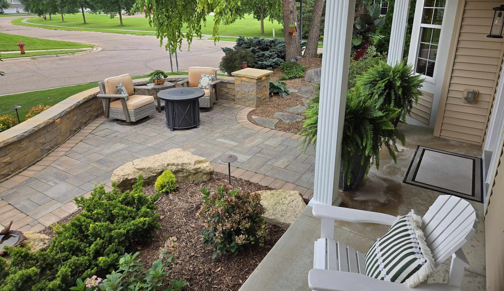 Patio with pavers, seating, fire pit, and landscaped areas. A house with porch and greenery.