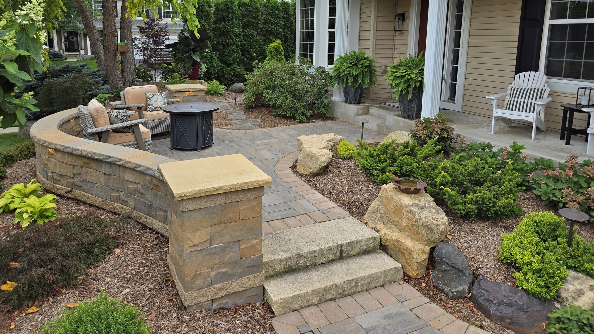 Stone patio with fire pit, seating, and landscaping leading to a house entrance.
