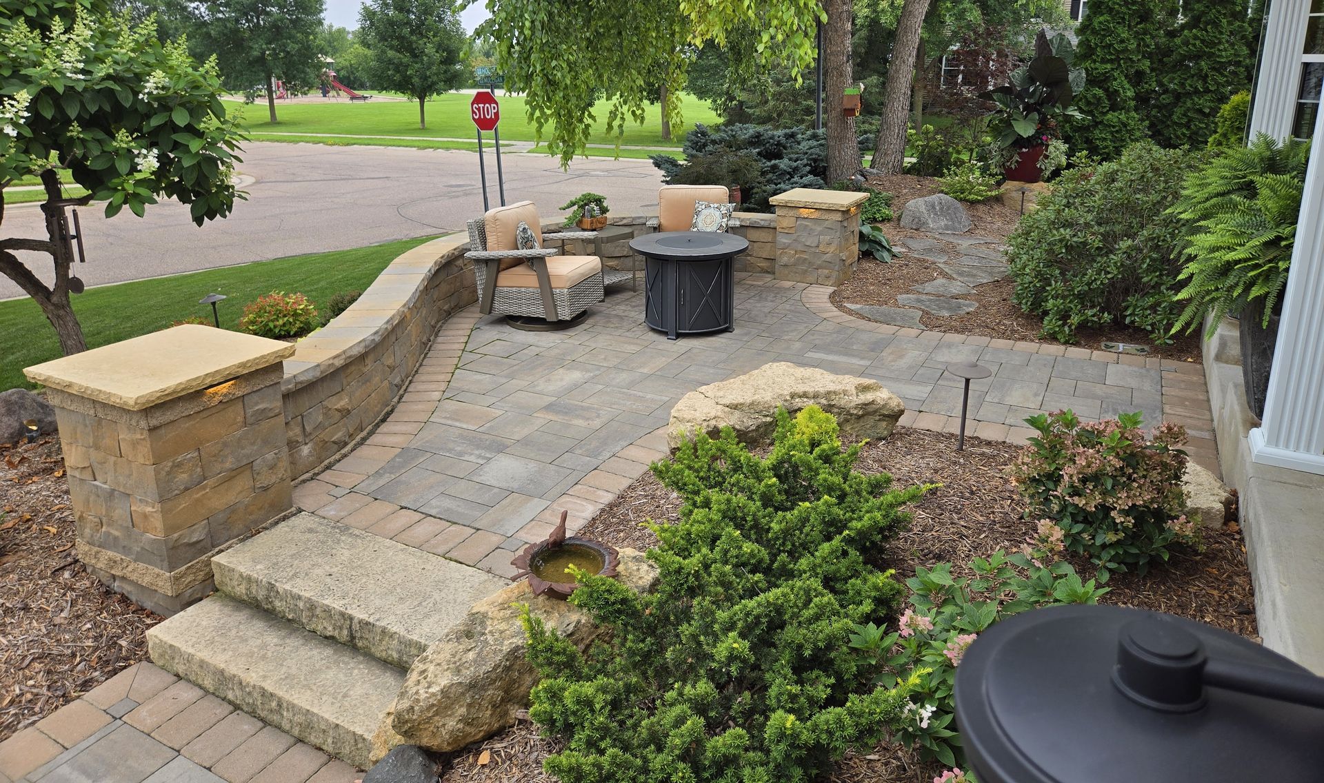 Stone patio with seating area, fire pit, and landscaped greenery near a residential street.