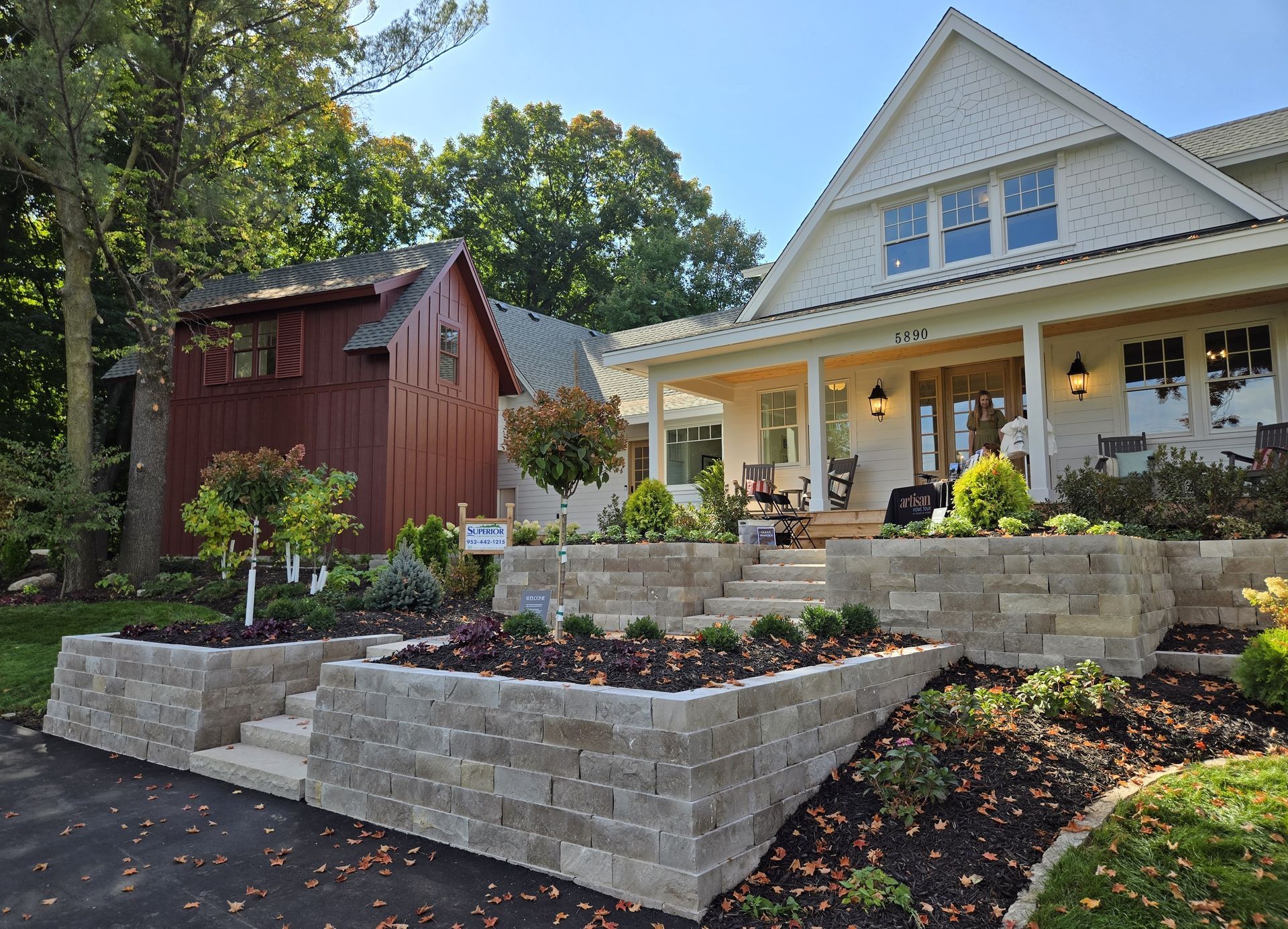 A two-story white house with a porch, terraced landscaping, and a dark red outbuilding under a blue sky.