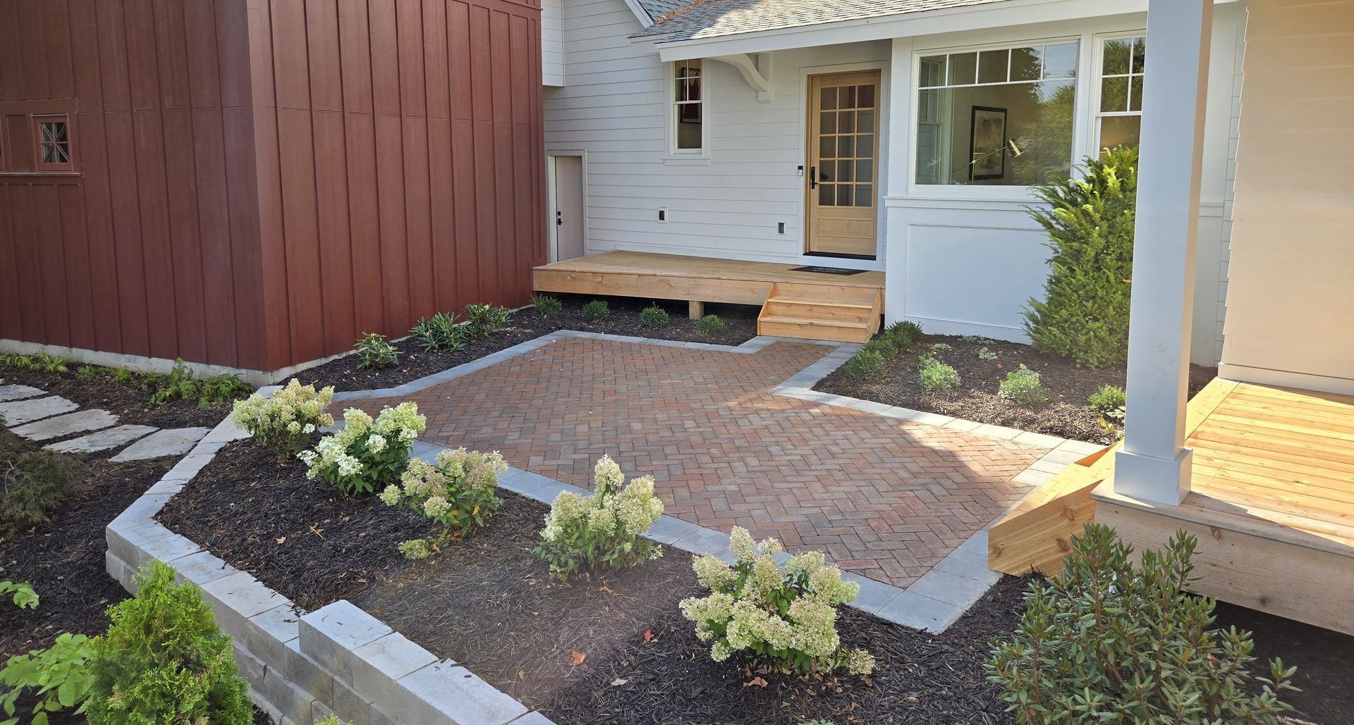 Brick patio with raised garden beds and wooden porch leading to a white house with a brown door.