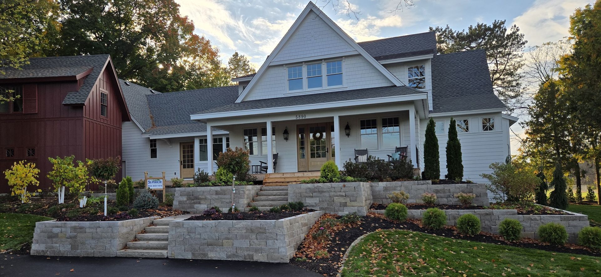 White farmhouse with stone retaining walls, front porch, and red barn on left.