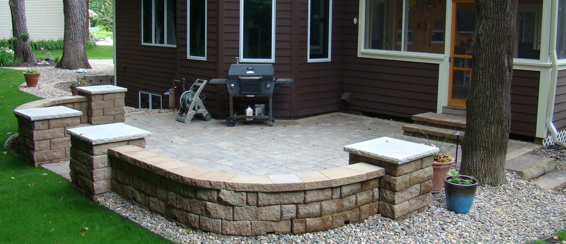 Stone patio with a grill and raised brick planters in a backyard.