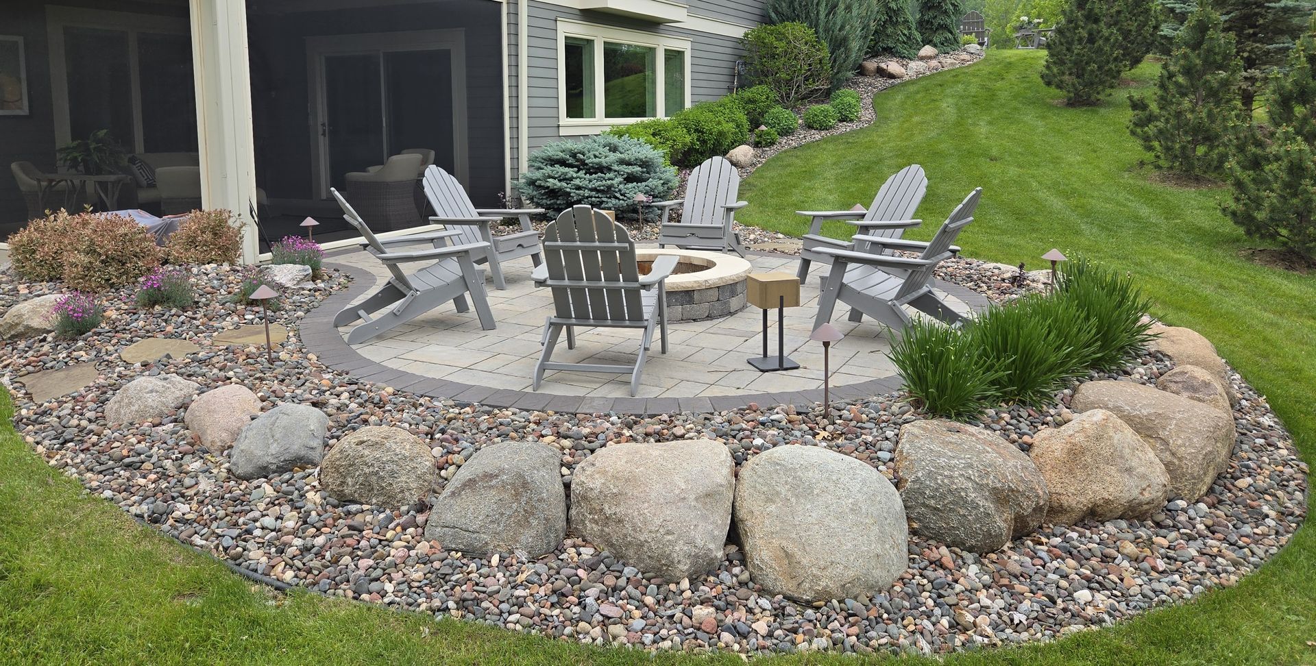 A backyard patio with Adirondack chairs around a fire pit, surrounded by rocks and landscaping.