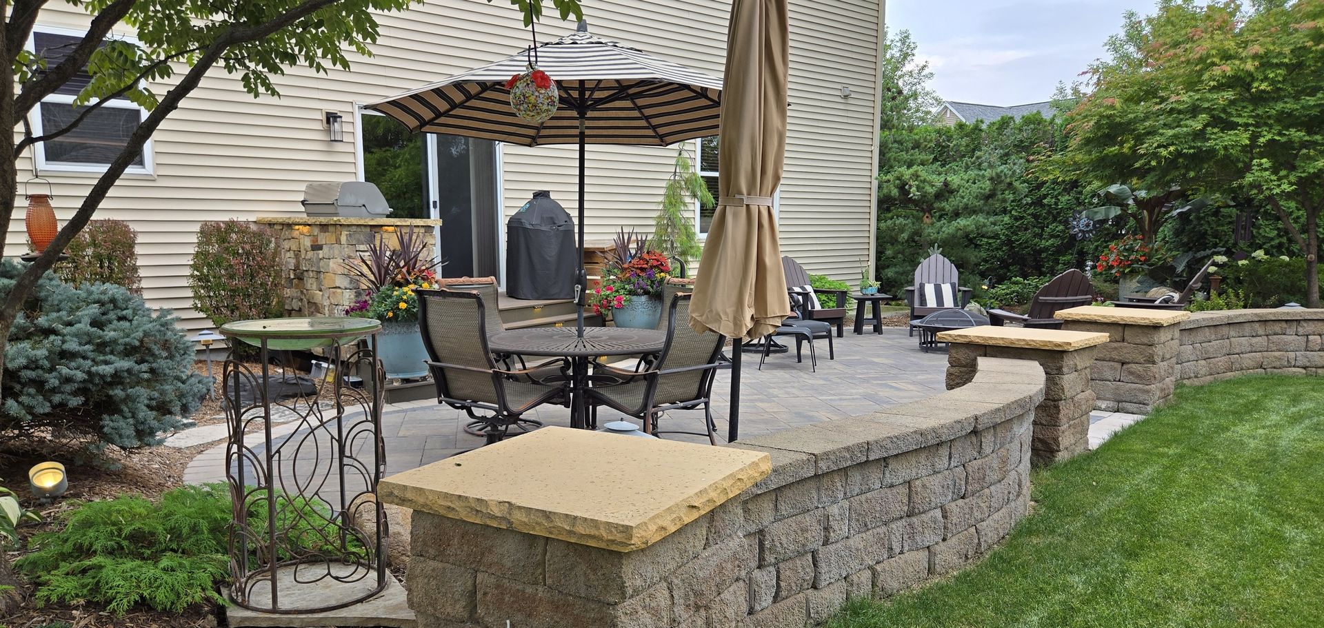 Patio with table, chairs, umbrella, and a retaining wall, surrounded by greenery.