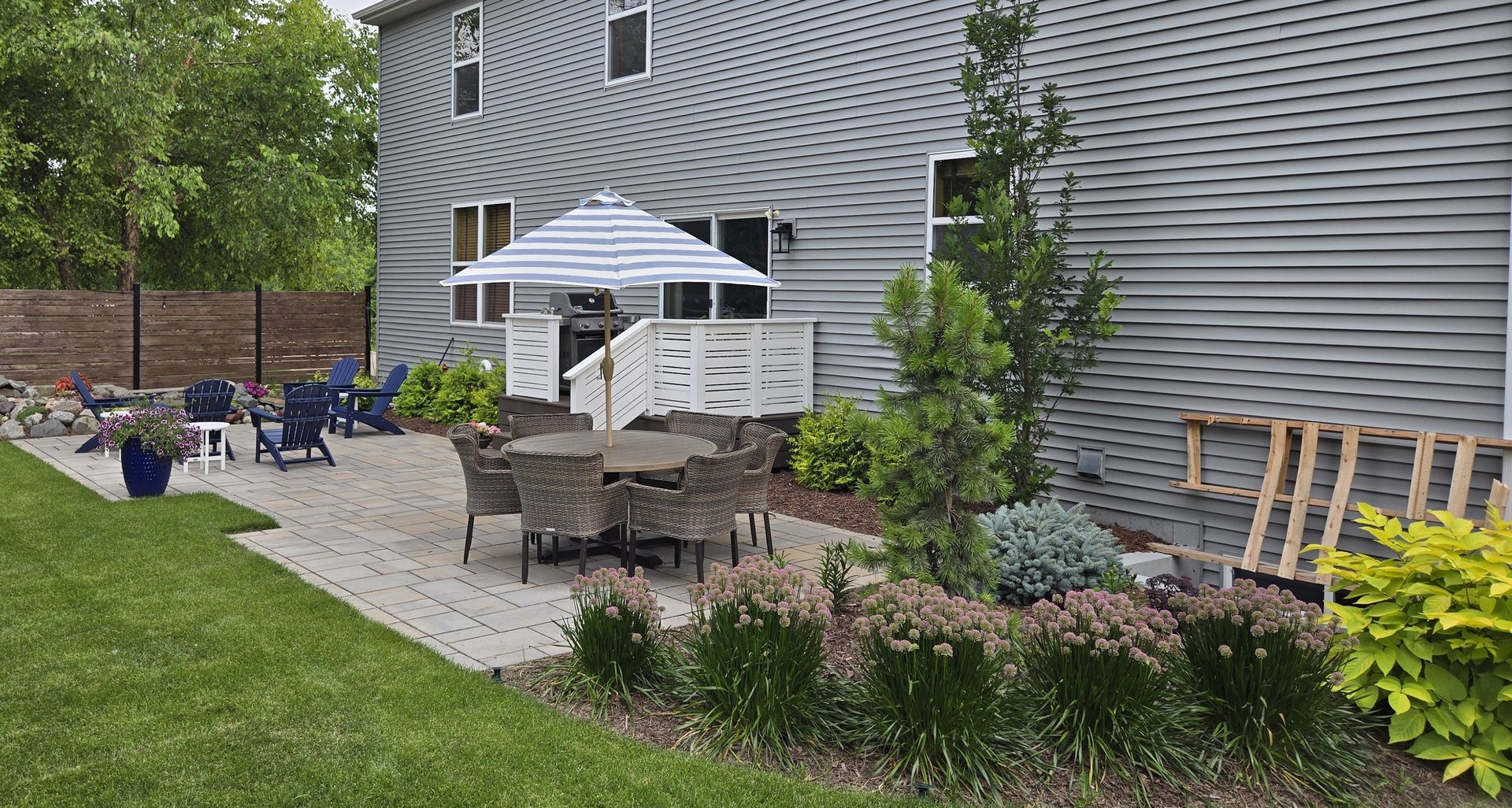 Backyard patio with dining set, umbrella, and landscaping next to a gray house.