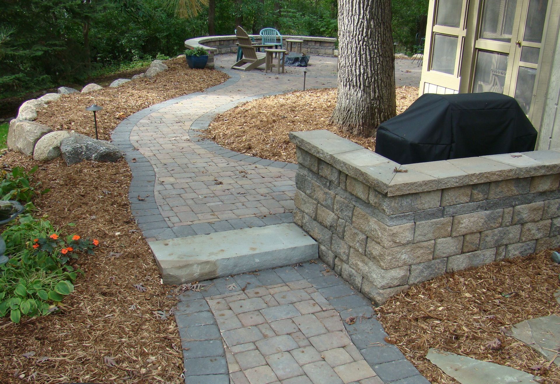 Brick pathway with mulch border, leading to a patio with outdoor furniture; grill to the right.